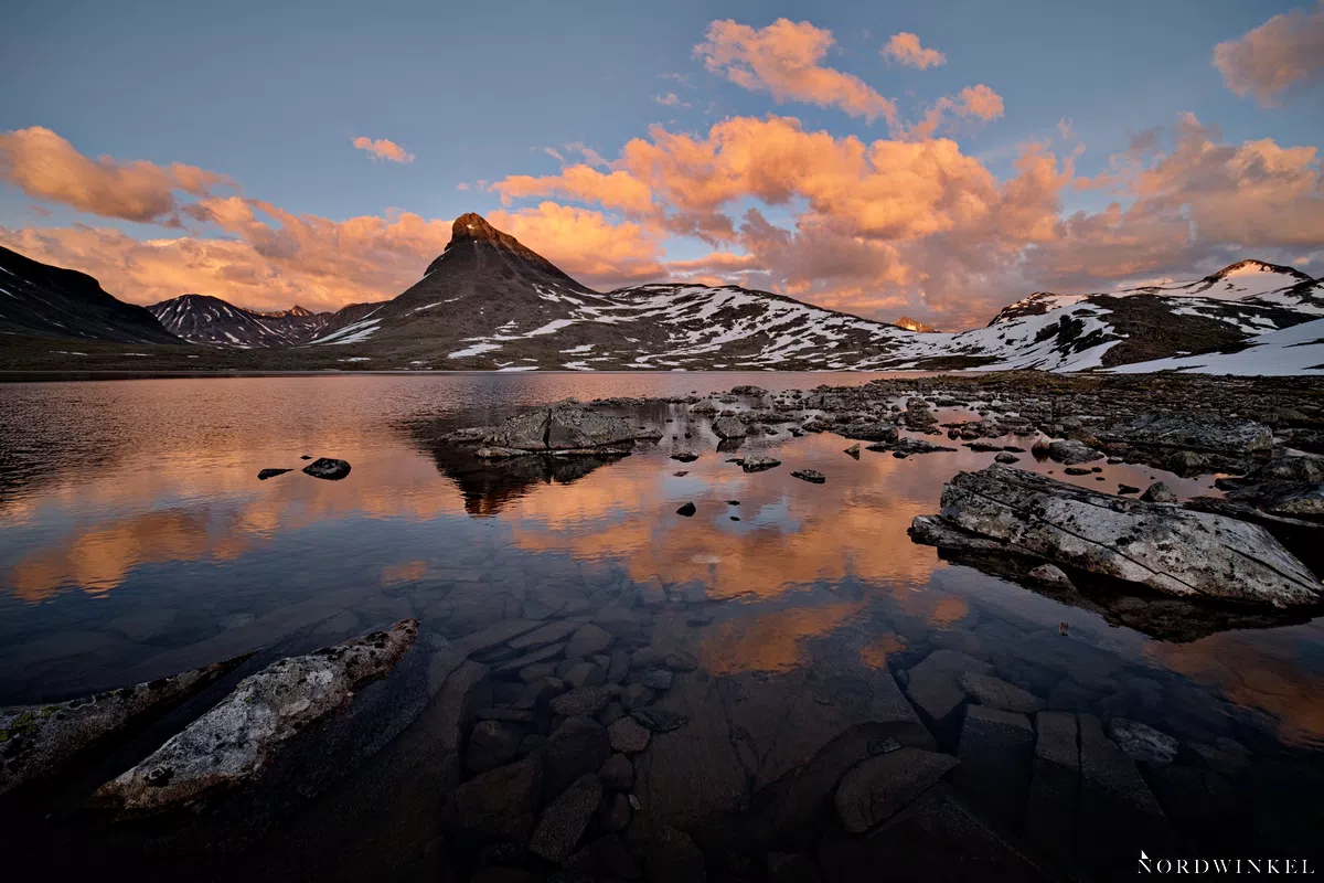 orange leuchtende wolken über schneebedeckten berggipfeln und einem see im vordergrund zeigt farbkontraste