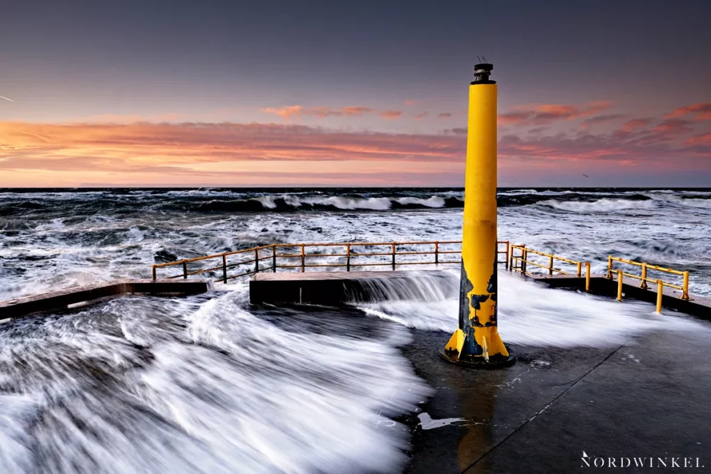 welle bricht über seebrücke mit gelber Leuchtbake zum sonnenuntergang