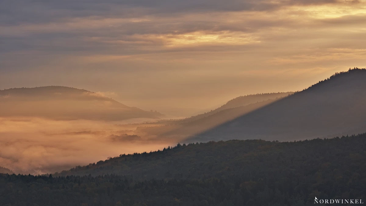 sonnenlicht bricht durch dichte wolkendecke und bestrahlt nebel zwischen bewaldeten bergen tiefenstaffelung sorgt für räumliche tiefe