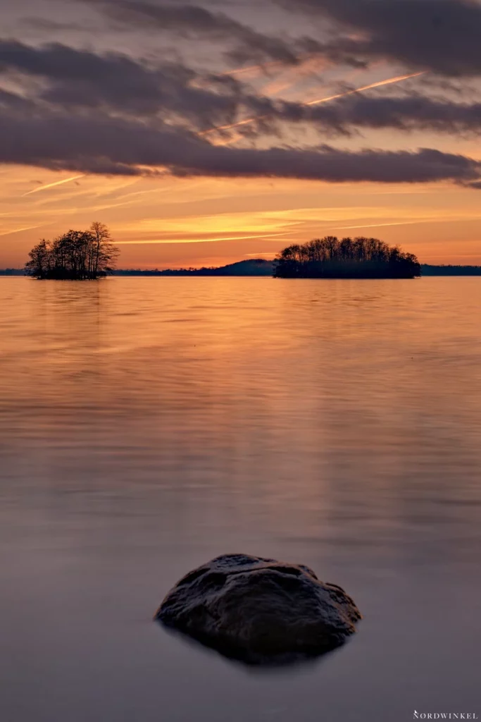 zwei inseln in einem see zum licht der untergehenden sonne mit einem stein im vordergrund für räumliche Tiefe