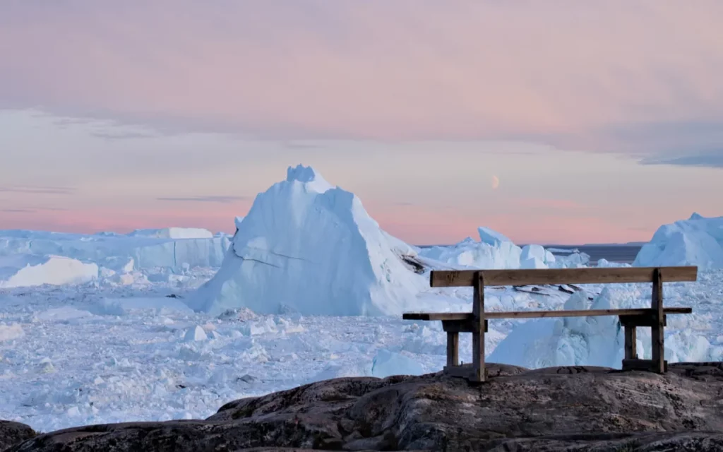 bank auf felsigem untergrund vor treibenden eisbergen in den farben des sonnenuntergangs